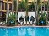 A swimming pool surrounded by lounge chairs, cabanas with white curtains, and tropical plants. The scene is set against a multi-story building backdrop with tall palm trees.