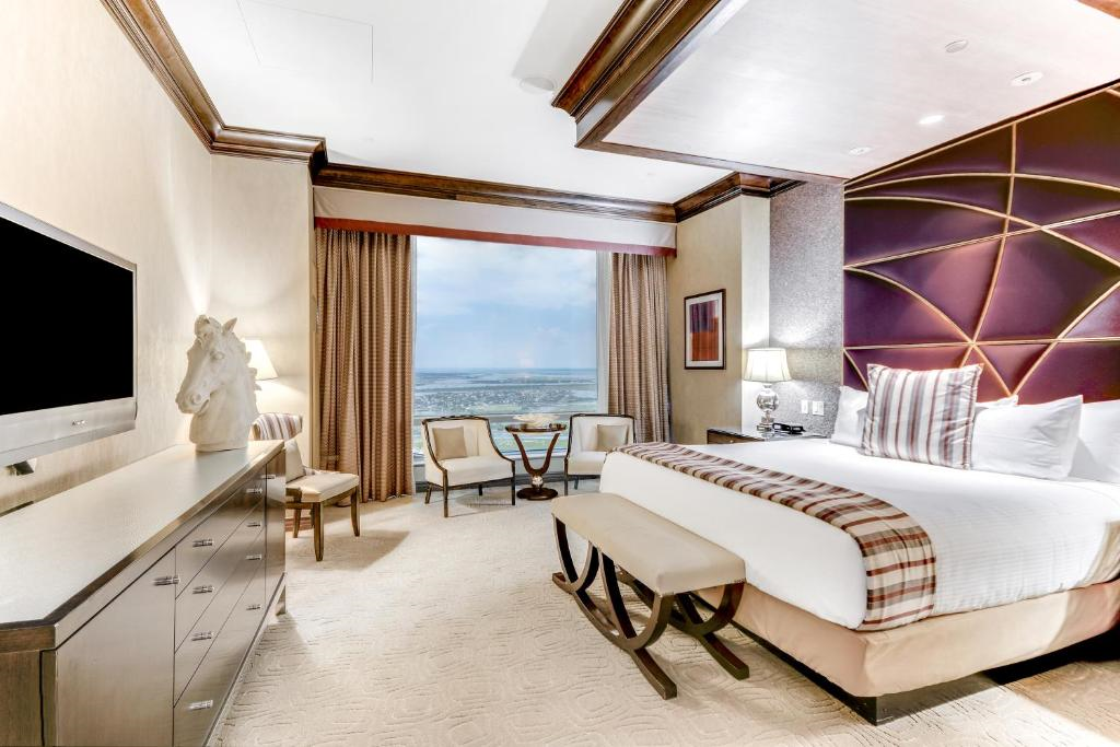 Elegant hotel room with a king-sized bed, modern decor, TV, and a window view of the ocean. A small seating area is positioned near the window with two chairs and a round table.