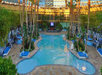 Indoor pool surrounded by palm trees and cabanas under a glass ceiling with blue lounge chairs and a TV screen at the far end.