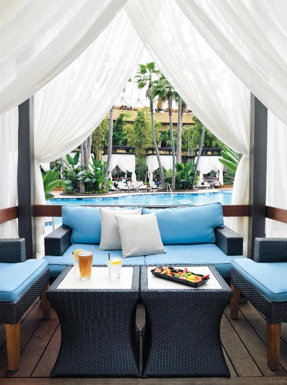 Outdoor cabana with blue seating, dark wicker tables, drinks, and a food platter. Pool and palm trees visible in the background, framed by white curtains.