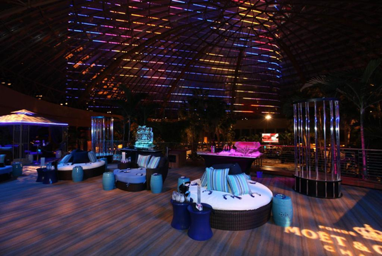 A dimly lit lounge with round seating, striped cushions, and colorful overhead lighting.