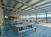 Large covered outdoor seating area with rows of wooden picnic tables and benches, overlooking water and trees in the background.