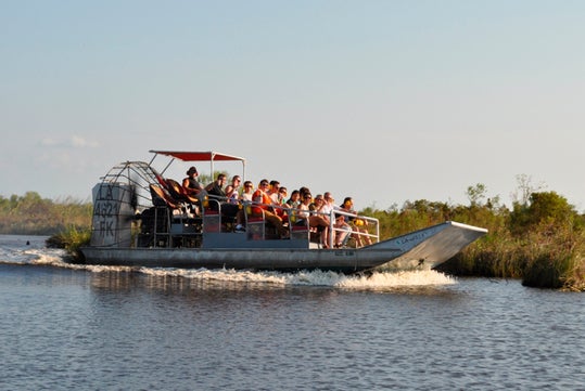 An airboat carrying passengers travels through a marshy waterway bordered by grass under a clear sky.