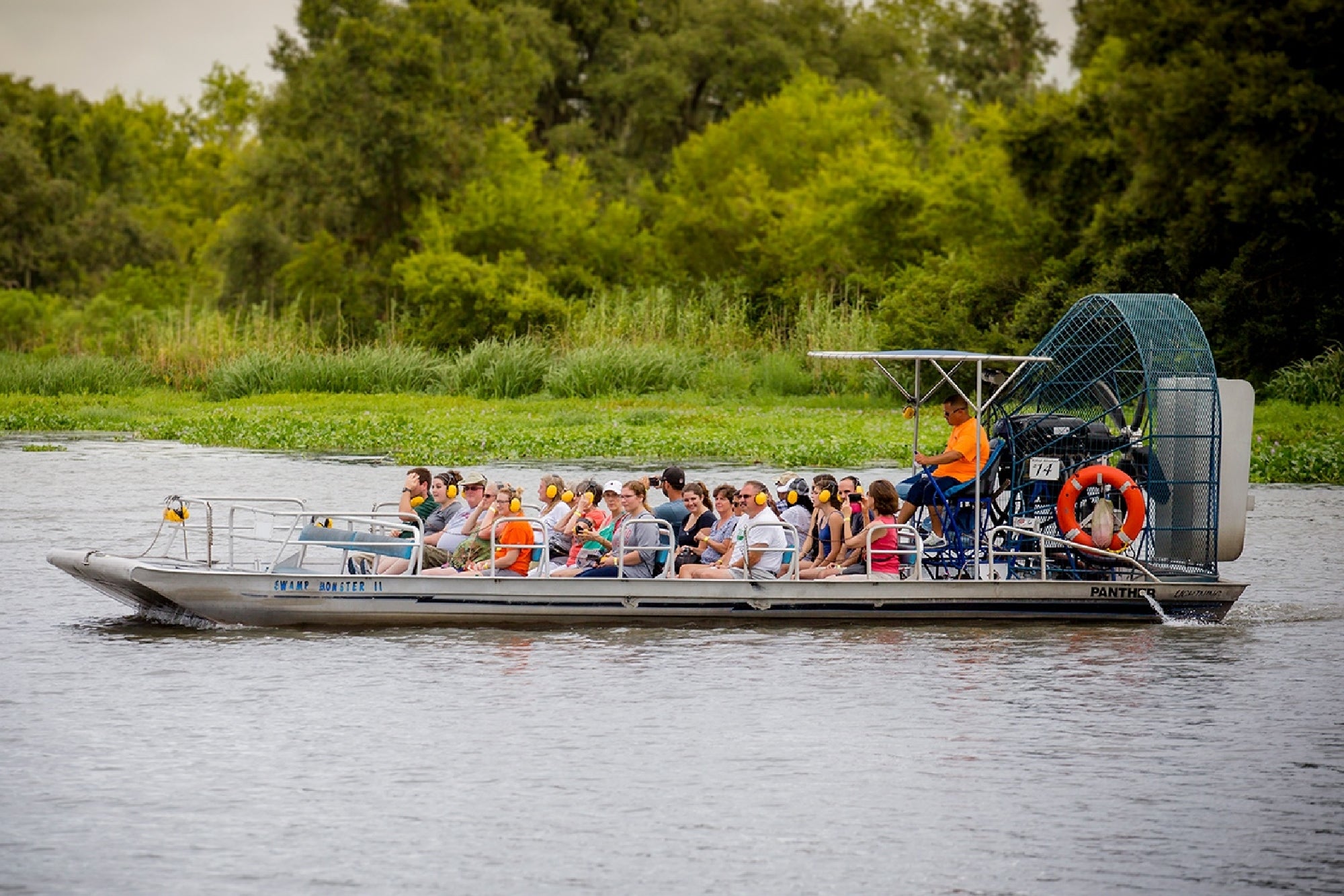 A group of people sit on an airboat with a large fan, traveling on a river with green vegetation and trees in the background.