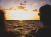 Two people watch the sun set over the ocean, with clouds scattered in the sky and golden light reflecting on the water’s surface.