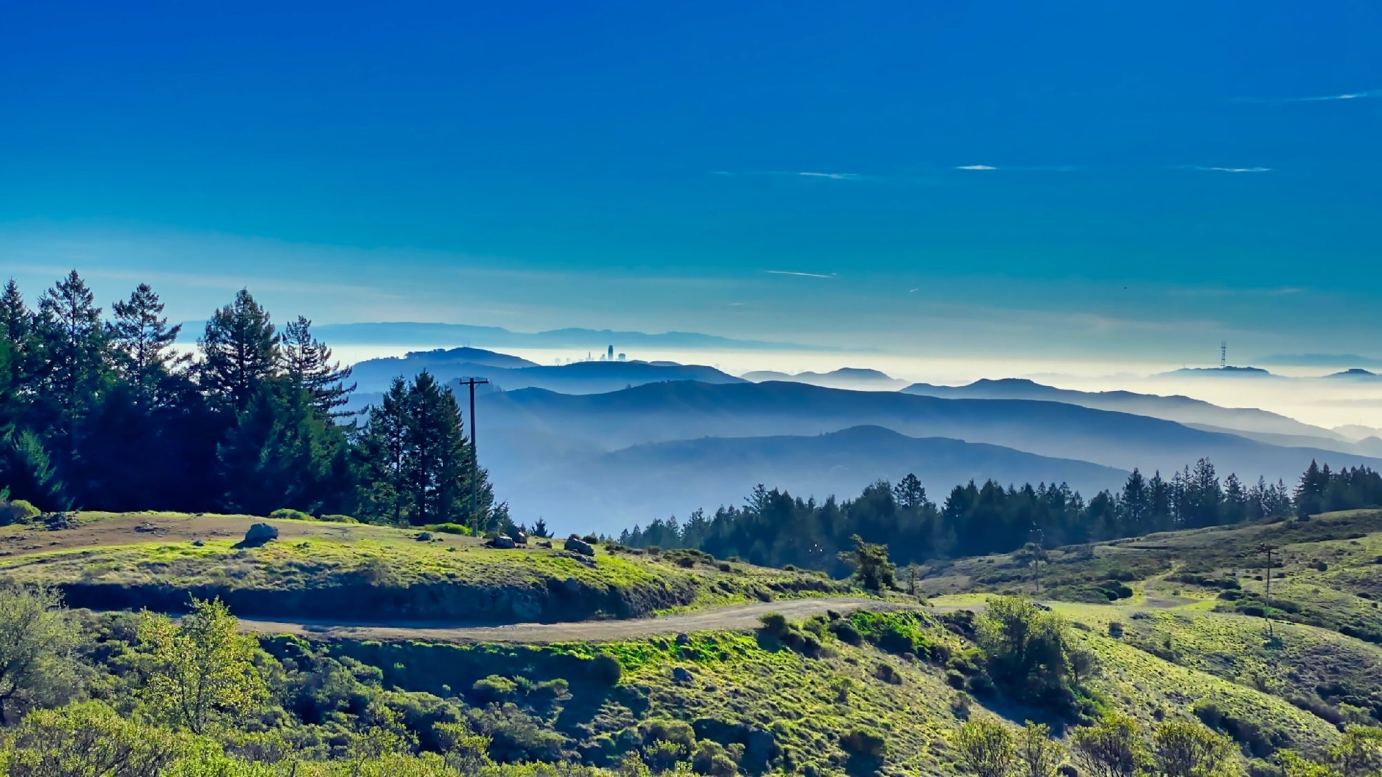 A scenic view of rolling green hills with a winding road and distant mountain peaks under a clear blue sky.