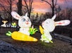 Two large, illuminated rabbit sculptures holding corn and a carrot are displayed outdoors at sunset, with more lighted animal figures in the background among bare trees.