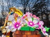 Illuminated lantern sculpture of a bearded man in overalls and hat standing beside oversized pink and white flowers, with bare trees in the background.