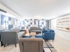 A modern lounge area with gray and blue sofas, a wooden coffee table, and a blue and white geometric accent wall. An abstract painting hangs on the right wall.