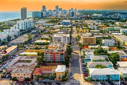 Aerial view of an urban area with numerous buildings and streets, featuring a mix of modern and older structures, with a city skyline and ocean in the background at sunset.