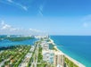 Aerial view of a coastal city with high-rise buildings along a sandy beach. The ocean is on the right, and the city extends toward the horizon under a clear blue sky.