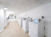 A clean, white laundry room with several washing machines and dryers lined up against the wall, under bright lighting.
