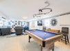 A spacious game room with a pool table in the foreground, seating area with sofas in the background, a TV on the wall, and modern lighting fixtures.