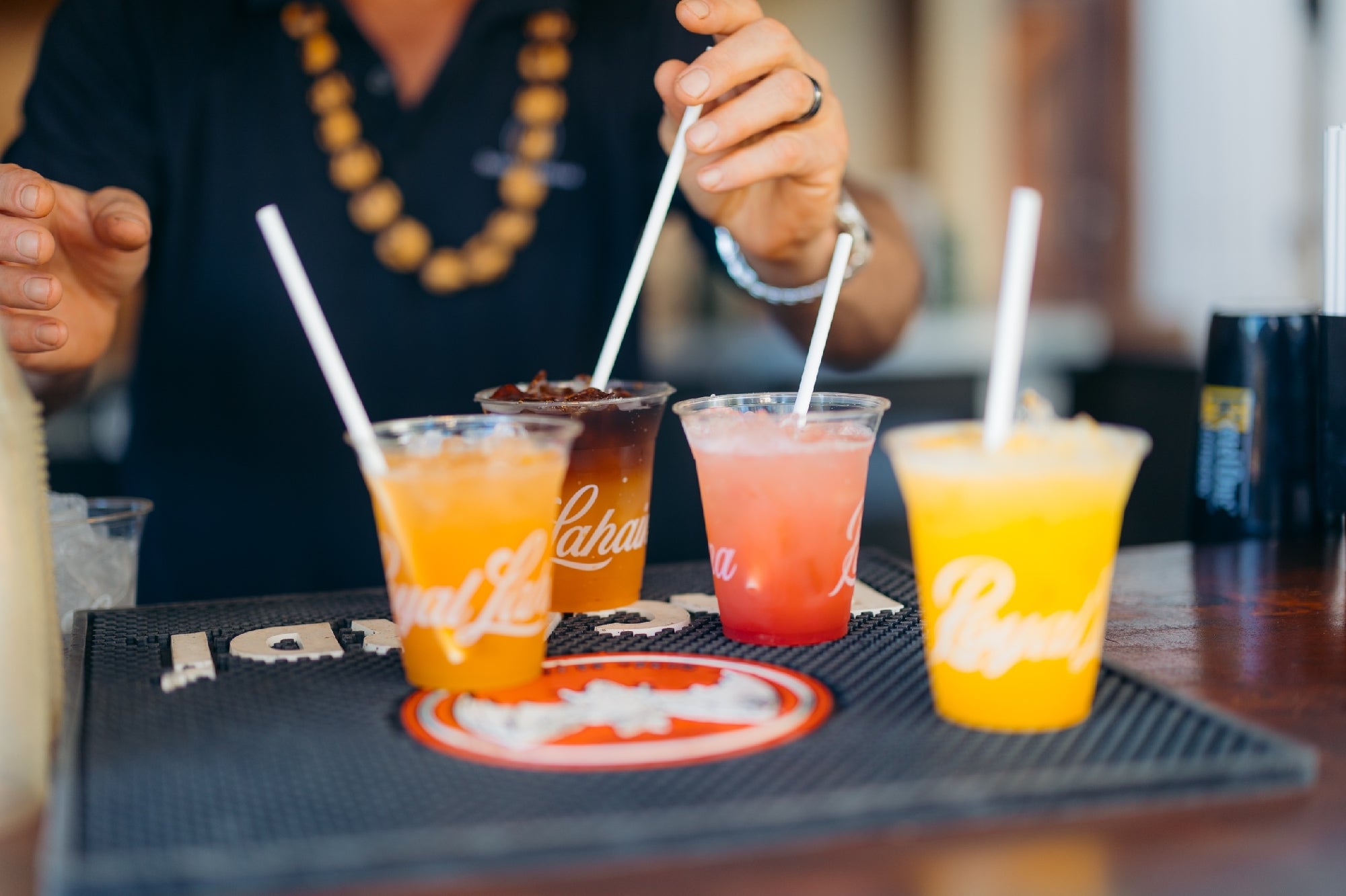 A person reaches for one of four colorful drinks with straws on a bar mat; the drinks are orange, pink, yellow, and dark brown.