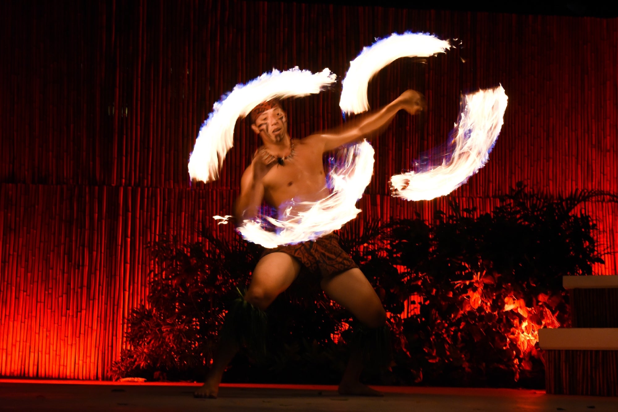 A performer in traditional attire spins two flaming batons on stage, creating arcs of fire against a backdrop of bamboo and red lighting.