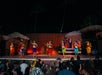 Six hula dancers and one male performer in traditional attire perform on an outdoor stage at night; audience members watch from tables decorated with flowers.