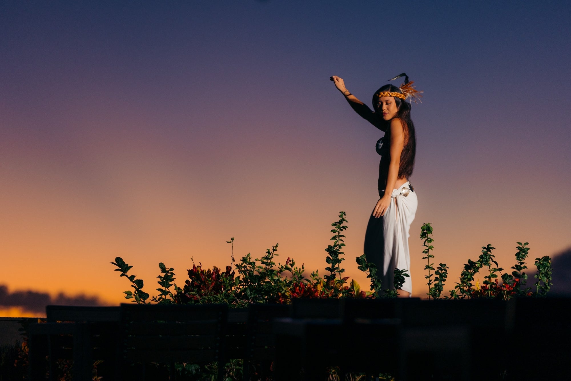 A woman in traditional attire performs a dance outdoors at sunset, surrounded by plants and silhouetted against the evening sky.