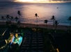 Aerial view of an outdoor event with a lit stage near a beach at sunset, featuring rows of seats, palm trees, and calm ocean water in the background.