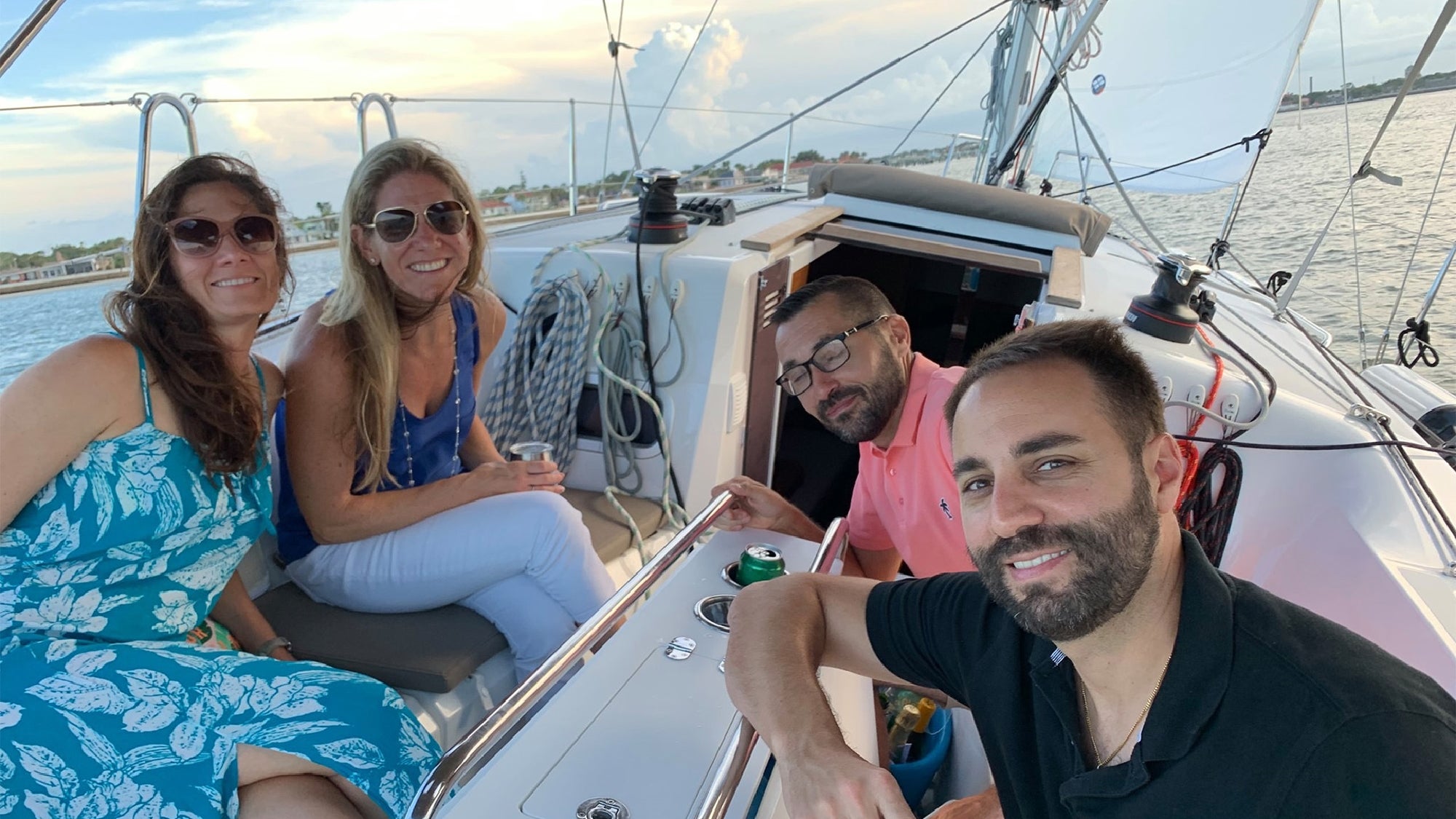 Four adults sit together on the deck of a sailboat, smiling at the camera with water and sky visible in the background.