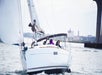 A white sailboat with people onboard sails on the water near a city shoreline, with a bridge visible on the right side of the image.