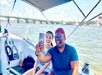 Two people sit on a boat, smiling as they take a selfie together on a sunny day, with a bridge and cityscape visible in the background.