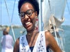 A woman wearing glasses and a sleeveless top smiles while standing on a boat, with water and a blurred figure in the background.