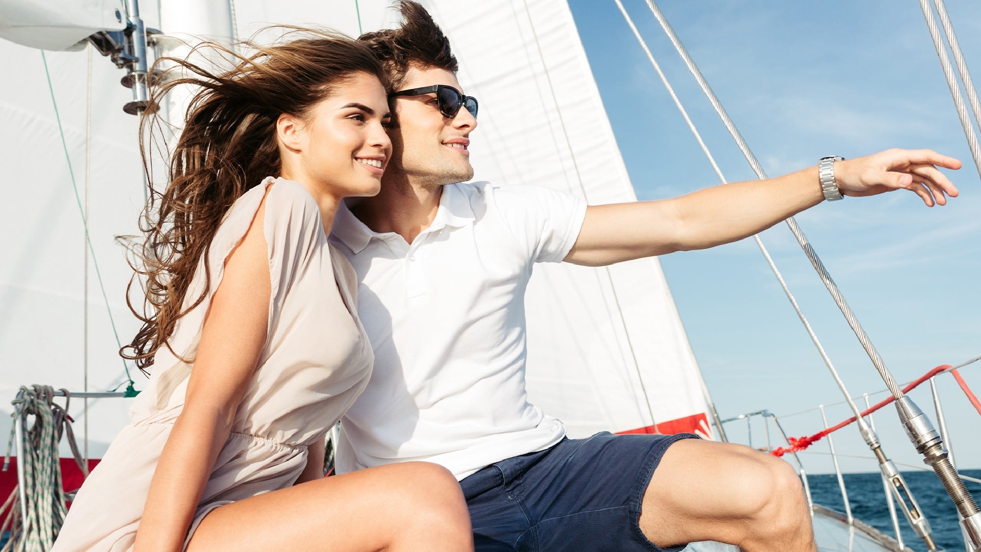 A young couple sits on the deck of a sailboat, smiling and looking out at the ocean under a clear blue sky. The man points into the distance.