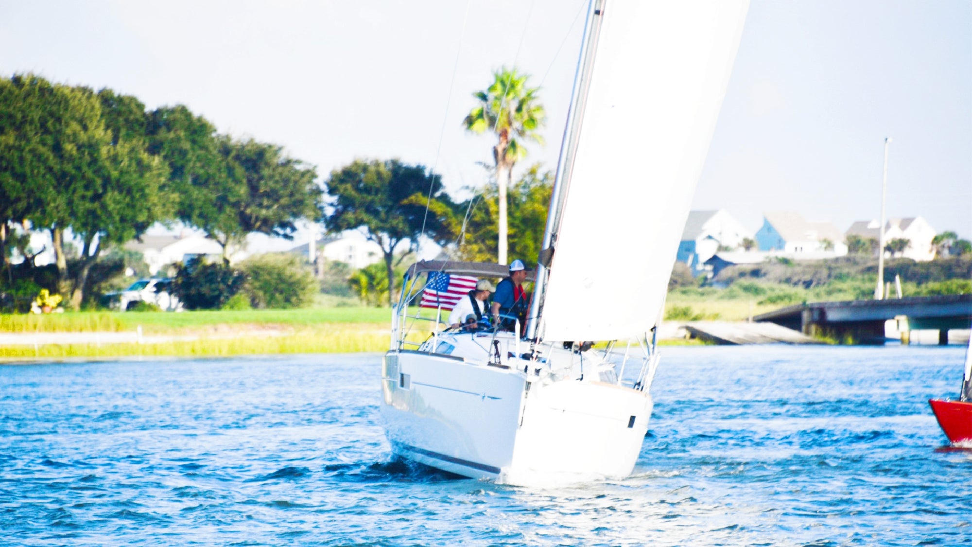 A white sailboat with an American flag sails on a calm waterway near green trees and houses.