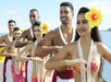 A group of dancers wearing colorful attire and leis perform a traditional dance by the sea.