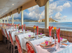 Dining area with tables set for a meal, featuring red napkins and floral decorations. Large windows offer a view of the ocean, city skyline, and a rainbow over a distant mountain.
