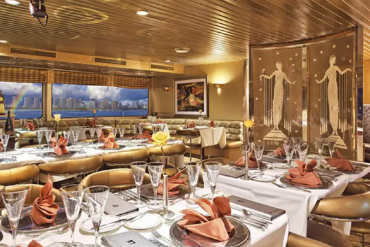 Elegant dining room with set tables, neatly folded napkins, and glasses. Large windows reveal a cityscape view. Decorative divider features artistic figures.