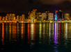 City skyline at night with tall buildings illuminated by colorful lights, reflecting on the water in the foreground.