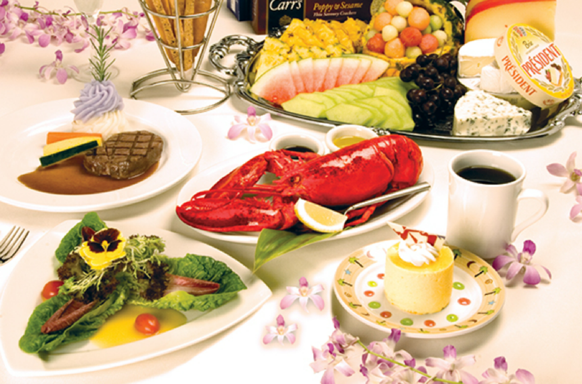 A luxurious meal with lobster, steak, colorful salads, assorted cheeses, fruits, coffee, and a small dessert on a white table accented with purple flowers.