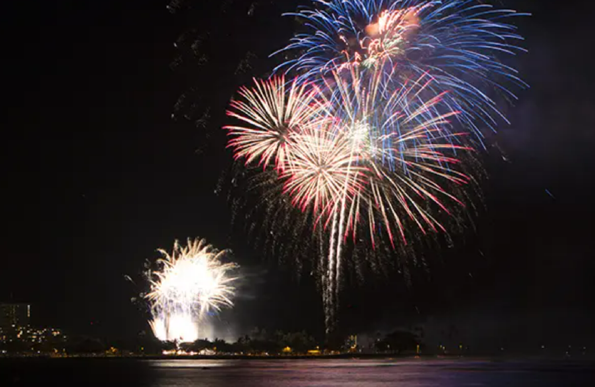 Colorful fireworks burst against the night sky over a body of water, with reflections visible on the surface.