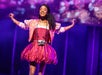 Person on stage smiling with arms outstretched, wearing a pink dress and jacket, with a camera around their neck. Background features a colorful light display.