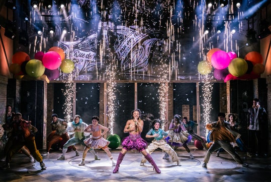 A lively stage performance with dancers in colorful costumes, bright lights, balloons, and fireworks effects, centered on a singer in a pink outfit.