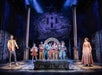 Actors perform on a stage set with four columns, a coffin center stage, and a painted backdrop resembling a mural; cast members wear colorful period costumes.