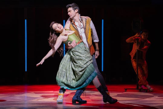 A man and a woman in costume perform a dramatic dance pose on stage, with a spotlight on them and another person in the background.