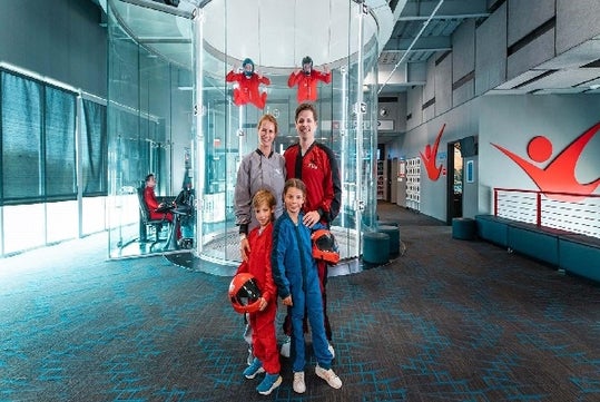 A family in jump suits standing in front of a flight tunnel with two instructors floating in the air behind at iFly.