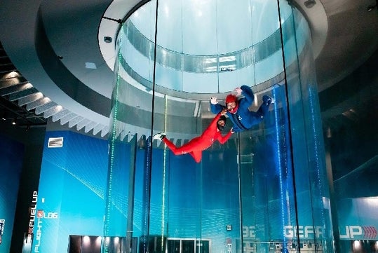 Two people in red and blue suits float inside a vertical wind tunnel at an indoor skydiving facility.
