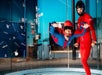 Person indoor skydiving in a wind tunnel, wearing a blue and red suit and helmet, assisted by an instructor in red gear.