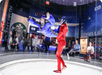 A person in flight gear floats inside a vertical wind tunnel while an instructor in a red suit observes; spectators watch through the glass wall.