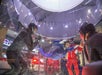A person in a blue suit is floating inside an indoor skydiving wind tunnel, surrounded by instructors in helmets and flight suits.