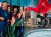 A group of people in flight suits watch and cheer as a person in a red suit and helmet does an upside-down indoor skydiving pose inside a vertical wind tunnel, high-fiving a girl through the glass.