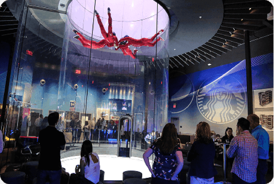 People watch two individuals in red suits performing maneuvers inside a vertical indoor skydiving wind tunnel at an entertainment facility.