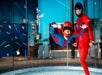 Two people in helmets and jumpsuits are inside an indoor skydiving wind tunnel; one is floating horizontally while the other stands nearby.