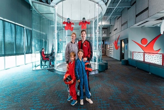 A family stands in front of an indoor skydiving wind tunnel while two people in red suits float inside. The environment is modern with glass walls and safety equipment visible.