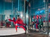 An instructor guides a person in a red suit inside an indoor skydiving wind tunnel while a group in helmets and suits observe from outside.