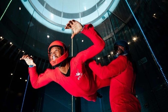 Two people in red suits indoor skydiving, one is airborne while the other assists.