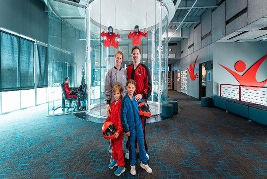 A family of four in flight suits poses with helmets inside an indoor skydiving facility; two instructors are floating in a vertical wind tunnel in the background.